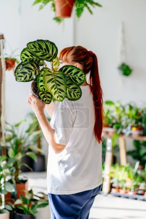 Téléchargez les photos : Style de vie durable, concept de vie lente. Jeune femme aux cheveux roux fleuriste, amateur de plantes, cacher son visage avec des plantes en pot dans la verdure magasin floral boutique. - en image libre de droit