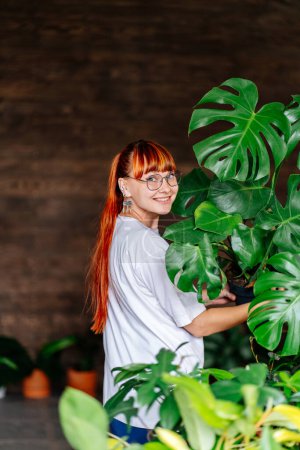 Téléchargez les photos : Plan vertical d'une jeune jardinière portant un t-shirt blanc, un jean bleu tenant une grande plante Monstera dans un pot en plastique. Jardinage à domicile, amour des plantes d'intérieur, freelance. concept - en image libre de droit