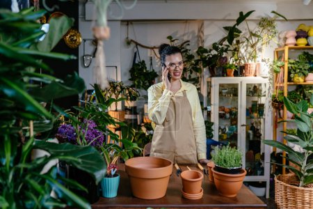 Photo for African american female owner or shop assistant wearing apron talking on mobile phone while working in plant store. - Royalty Free Image