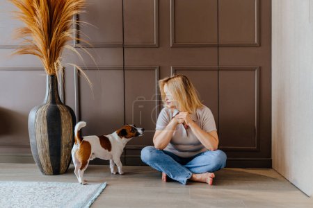 Foto de Actividad en el hogar, concepto de tiempo libre. Una mujer madura con su gato Russell terrier están sentados en el suelo. La mujer habla con el perro y lo mira. - Imagen libre de derechos