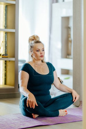 Photo for Vertical shot of full-figured mature middle age woman sitting on mat in lotus pose. Calm lady practicing techniques and yoga indoors. - Royalty Free Image