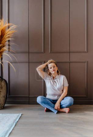Téléchargez les photos : Portrait vertical de charmante femme blonde mature élégante assise sur le sol à la maison. - en image libre de droit