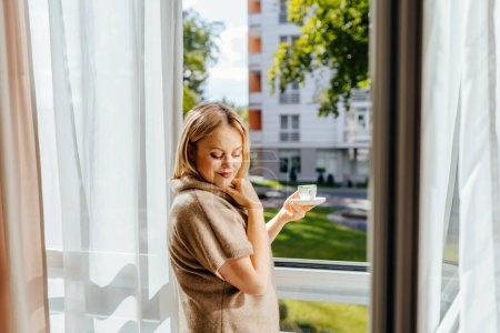 Téléchargez les photos : Charmante belle femme blonde mature de 55 ans debout devant la fenêtre sous les rayons du soleil, buvant du café ou du thé le matin, rêvant d'imaginer leur avenir. - en image libre de droit