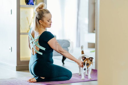 Foto de Una mujer rubia encantadora madura de tamaño grande se sienta en la esterilla de yoga juega con su perro mientras hace ejercicios deportivos en casa con ropa deportiva. - Imagen libre de derechos