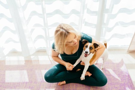 Photo for A plus-size mature charming blond woman sit at Pilates mat hugging with her dog while making sports exercises at home in sports clothes. - Royalty Free Image