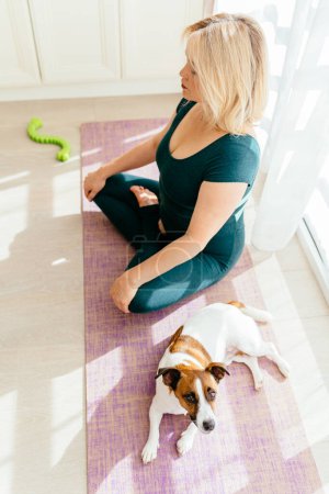 Téléchargez les photos : Plan vertical de femme mature blonde 55s pratiquant l'exercice avec le chien pour profiter et se détendre avec le yoga. Accueil d'entraînement sportif avec animaux domestiques. - en image libre de droit