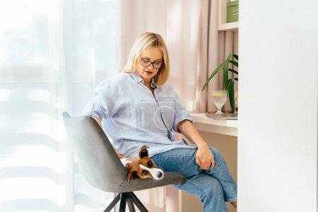 Foto de Mujer de negocios acariciando con portátil en casa perro sentado en la silla. Una mujer madura activa con un perro que trabaja en la oficina en casa, mirando y utilizando el ordenador portátil. - Imagen libre de derechos