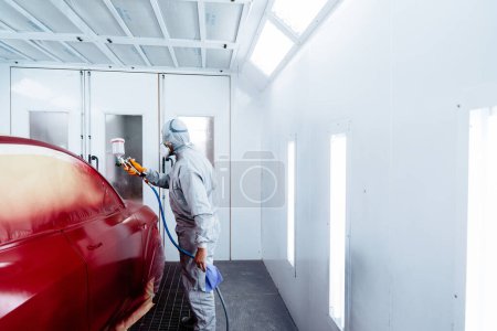 Téléchargez les photos : Technicien en peinture masculine appliquant de la peinture rouge sur une pièce de voiture pistolet pulvérisateur dans un atelier de carrosserie. - en image libre de droit