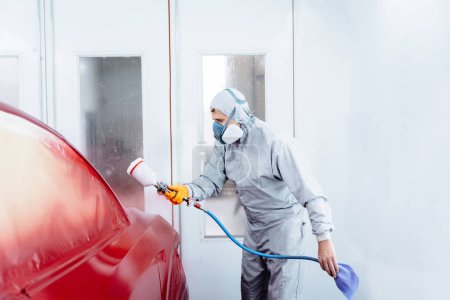 Photo for Male worker in grey costume cloth wearing chemical protective mask using spray gun and airbrush and painting a red car. - Royalty Free Image