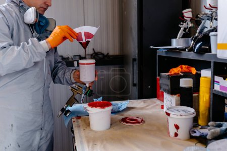 Männlicher Autolackierer in Schutzkleidung schüttet an einer Tankstelle gemischte Farbe aus einer Dose in einen Plastikeimer.