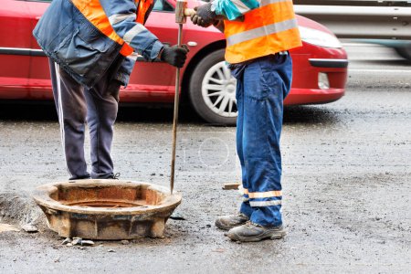 Photo for Road workers in orange overalls are repairing an old sewer manhole on the roadway against a blurred background of a passing car. Copy space. - Royalty Free Image