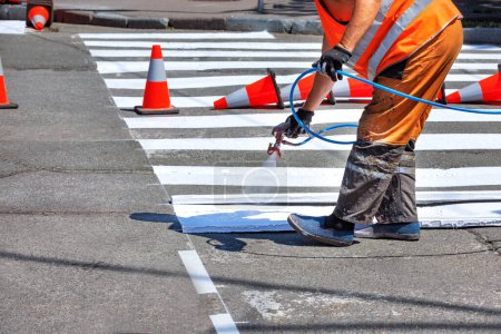 A road worker applies white road markings to a crosswalk using an airbrush and a wooden template surrounded by traffic cones.