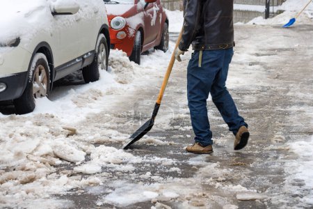 A man cleans snow and ice with a shovel from an asphalt path near parked cars in the courtyard of the house.