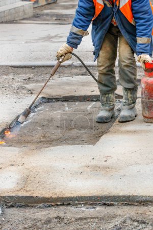 Photo for Pothole repair of the road. A worker using a gas burner and a propane tank heats the asphalt pavement for better bitumen adhesion during re-paving. Vertical image. Copy space. - Royalty Free Image