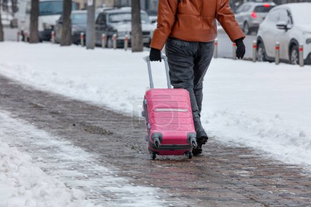 Foto de A woman in a brown jacket and gray trousers carries a bright pink travel bag on wheels along the cobbled sidewalk against the backdrop of a winter city street in blur. Copy space. - Imagen libre de derechos