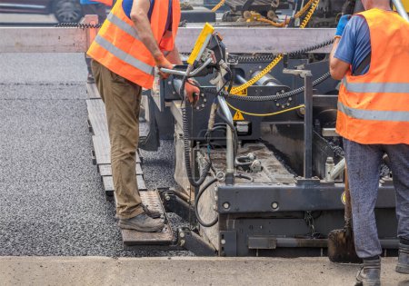 Foto de Road specialists in orange vests are laying fresh asphalt with a paver on the carriageway. Copy space. - Imagen libre de derechos