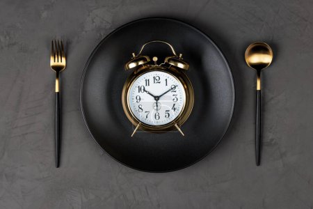 Foto de Gold-rimmed alarm clock, black plate and gilded cutlery fork and spoon on a dark gray concrete background. Flat lay. Diet regime. Copy space. - Imagen libre de derechos