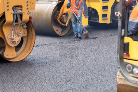 Photo for A road worker uses a shovel to level a layer of fresh asphalt against a background of road rollers. Copy space. - Royalty Free Image