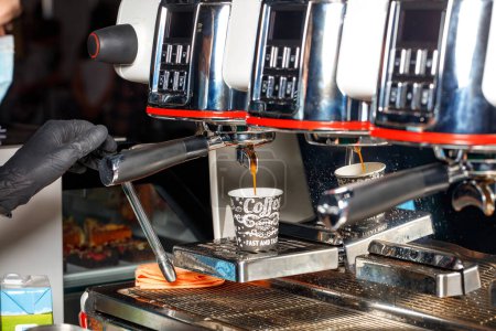 Foto de La mano de un barista en un guante de látex negro vierte café expreso caliente en una taza de papel de una máquina de café. - Imagen libre de derechos