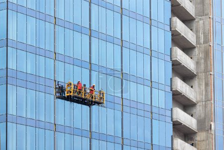 Foto de Acristalamiento por un equipo de trabajadores de la fachada de un nuevo rascacielos en un moderno edificio residencial utilizando una plataforma suspendida. Sellado, trabajos de fachada limítrofes. - Imagen libre de derechos
