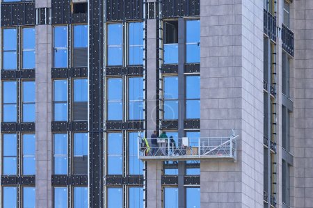 Une équipe de travailleurs sur un berceau suspendu effectue des travaux de façade de bordure dans un bâtiment résidentiel moderne en utilisant une plate-forme suspendue.