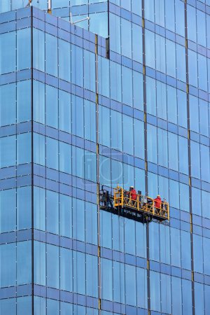 Bauarbeiter in roten Overalls führen die Verglasung der Fassade eines neuen Hauses in einer modernen Wohnanlage über eine schwebende Plattform durch. Abdichtungs- und Fassadenarbeiten. Kopierraum. Vertikales Bild.