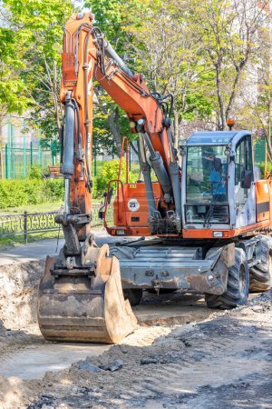 Photo for A large wheeled excavator is parked in a trench he dug in the middle of a city street road on a bright sunny spring day. Vertical image. - Royalty Free Image