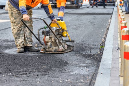Photo for A road worker repairs a section of road by compacting fresh asphalt with a vibrating plate around a manhole. Copy space. - Royalty Free Image