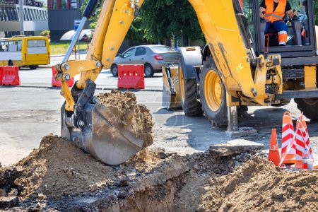 Un operador de excavadoras de construcción opera hábilmente un auge y un cubo para cavar una trinchera y descargar arena en un sitio de trabajo en el contexto de una calle de la ciudad en un día soleado de verano. Copiar espacio.