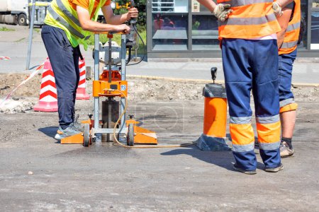 Road workers in reflective capes drill asphalt with a core drilling machine to take and measure cores during road repairs on a sunny day. Copy space.