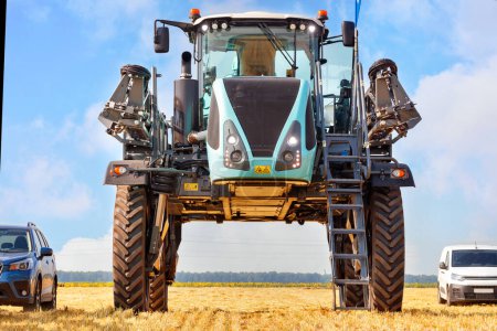 Photo for A high wheelbase industrial sprayer for the agricultural sector stands in comparison with passenger cars in a harvested field against a blue cloudy sky. - Royalty Free Image