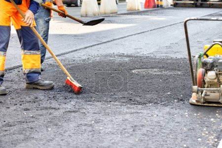 Photo for A team of road workers in orange overalls use a broom and shovel to level the fresh asphalt to evenly distribute the pavement around the sewer manhole before ramming it. Copy space. - Royalty Free Image