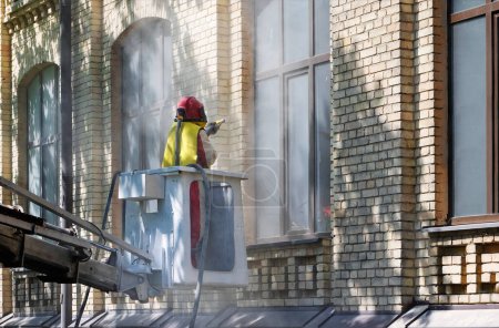 Ein Arbeiter im Schutzanzug, der in einer Konstruktionsteleskopwiege steht, reinigt mit einem Sandstrahler den verschmutzten Belag von der Ziegelwand der Hausfassade..