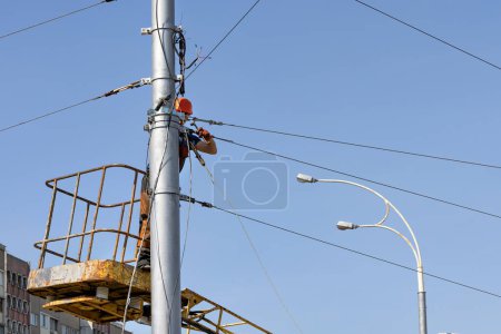 Photo for An electrician worker works on a light pole for a power line while standing in a lifting construction basket and securing a metal cable into a tripwire. Copy space. - Royalty Free Image