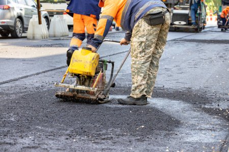 Photo for A road service worker compacts fresh asphalt on a section of road with a worn-out gasoline vibrating plate around a sewer manhole. Copy space. - Royalty Free Image