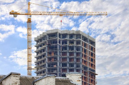 A tower crane works on the construction site of a modern multi-storey residential building against the background of a blue slightly cloudy sky, copy space.