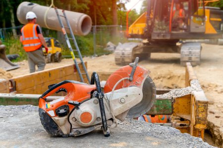 Photo for Orange petrol saw with a diamond blade for cutting concrete against a blurred background of drainage works on construction site - Royalty Free Image