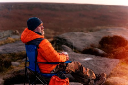 Bärtiger Mann in orangefarbener Jacke entspannt sich allein auf dem Gipfel des Berges und trinkt bei Sonnenaufgang heißen Kaffee. Reiselebensstilkonzept Der Nationalpark Peak District in England