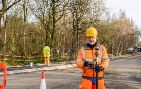 Photo for Site engineer operating his touch screen controller instrument during roadworks. Builder using touch screen controller to control total positioning station tachymeter on construction site for new road setting out - Royalty Free Image