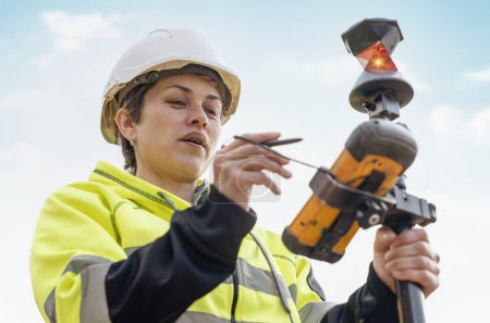 Téléchargez les photos : Portrait en gros plan d'une femme ingénieur de chantier qui travaille avec l'équipement EDM de la station totale de théodolite sur un chantier de construction à l'extérieur - en image libre de droit