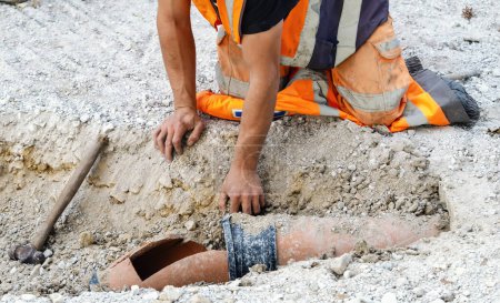 Builder in orange hi-viz clothing hand diggin and exposing damaged drainage pipe for repair
