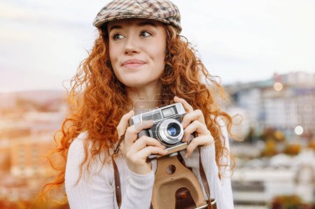 Photo for Enthusiastic readhead female model in classic English cap laughs happily enjoys spare time taking photo with old fashion camera outside against blurred city background. Lifestyle concept - Royalty Free Image