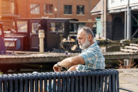 a bearded man senior talking by phone on the quays, pier, sitting on bench, looking at yacht, sailboat, boat. Lifestyle concept