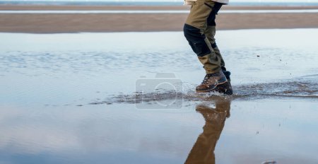 An energetic man in waterproof hiking boots walking along sea shore through water and puddles making splashes