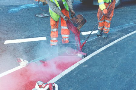 Téléchargez les photos : Travailleurs de la route appliquant la peinture rouge chaud marquage routier sur la nouvelle route de construction - en image libre de droit
