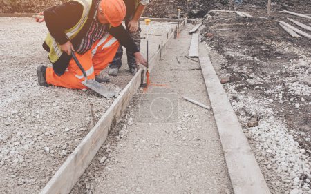 Photo for Groundworker in orange safety hi-vis trousers fixing a timber along string line with a steel pin to form a kerb riser and a straight edge for tarmac road surface during new road construction - Royalty Free Image
