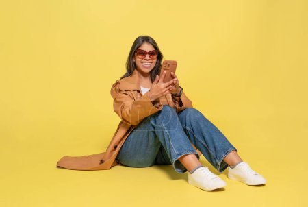mujer en jeans, una camiseta blanca y un abrigo marrón usando un teléfono móvil sentado sobre un fondo amarillo. Gente feliz