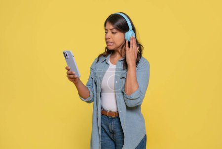 Unhappy Indian woman looking at her phone, wearing stereo headphones on her ears, having, reading bad news, dressed in jeans and denim shirt isolated on  a yellow background