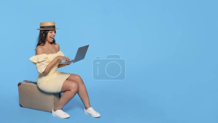 Mujer india en vestido amarillo sentada en la maleta usando portátil sobre fondo azul. Viajes disfrutar concepto
