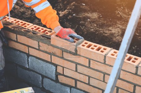 Industrial bricklayer laying bricks on cement mix on construction site. Fighting housing crisis by building more affordable houses concept